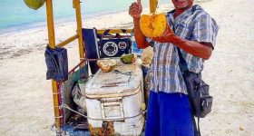 Homem numa barraca a vender cocos em Holbox, com ambiente tropical e praia ao fundo.